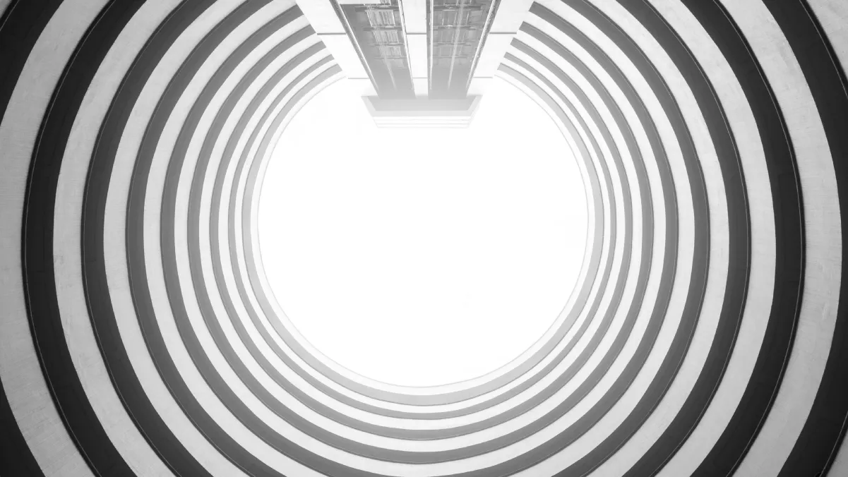 A view looking up into a circular architectural structure featuring concentric black and white stripes, leading to a bright, white center. The image conveys a sense of depth and perspective, with the walls curving inward toward the light.