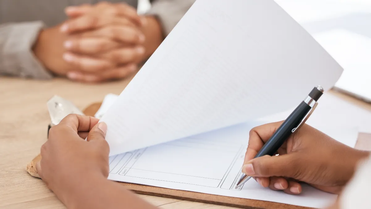 A person is holding a pen and writing on a blank sheet of paper, with their other hand resting on the table. In the background, another person's hands are clasped together, indicating a conversation or meeting taking place. The setting appears to be a professional or office environment.