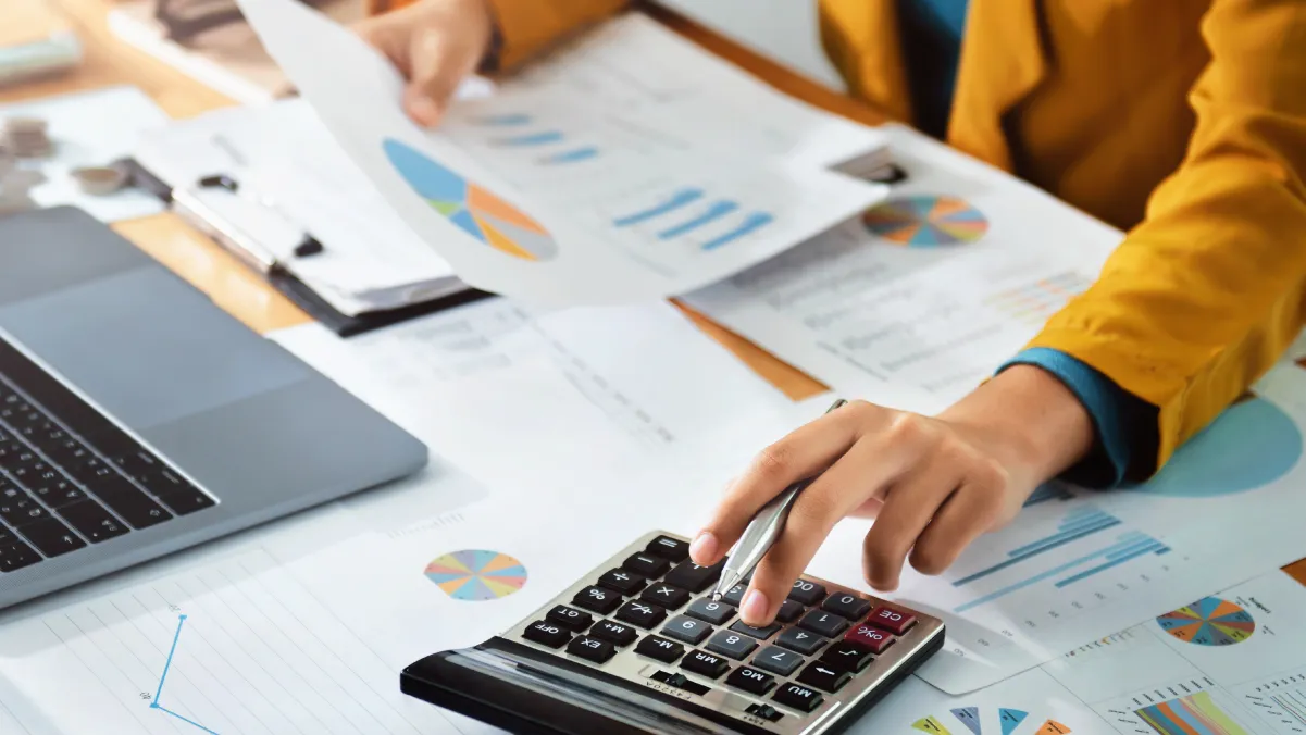 Person working on calculator with financial charts and laptop on desk.
