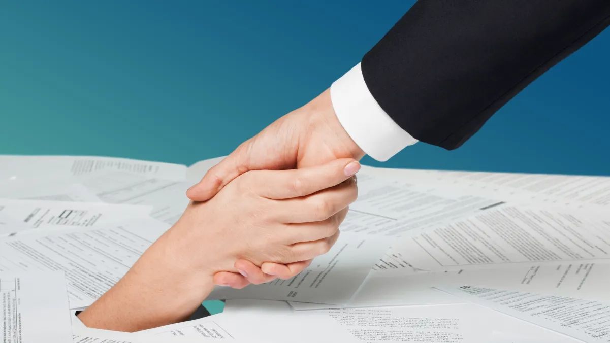 Two people shaking hands over documents with a blue backdrop.