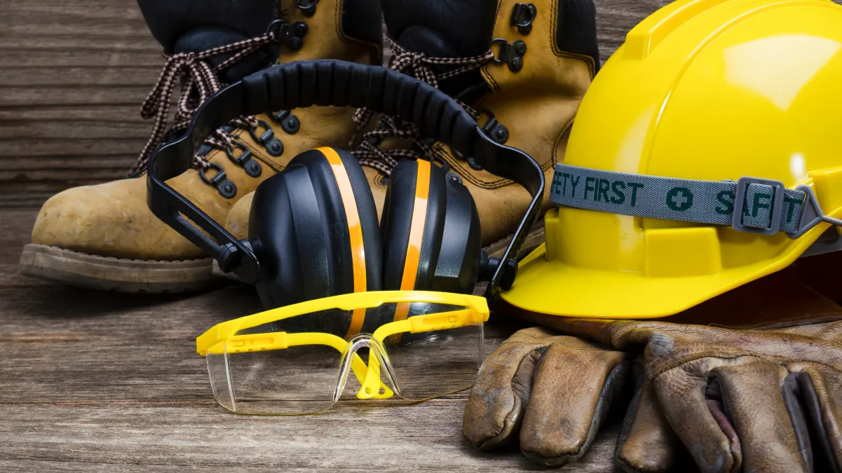 Work boots, ear defenders, safety glasses, hard hat, and gloves on a wooden surface.