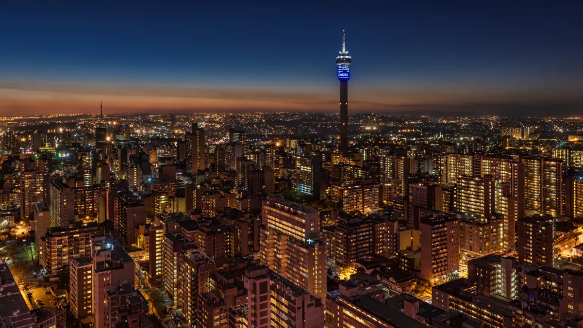 A panoramic night view of a vibrant city skyline illuminated by numerous lights, with tall buildings and structures visible. The iconic tower, likely a communication or observation tower, stands prominently in the center, glowing with blue lights. The scene captures the transition from dusk to night, showcasing a gradient sky that shifts from orange to deep blue. The city's layout reveals a mix of modern architecture and urban density, emphasizing the bustling nature of the area.