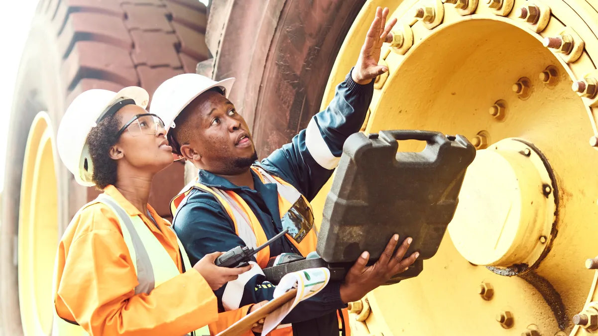 Two workers wearing hard hats and hi-vis vests inspecting machinery with a clipboard.